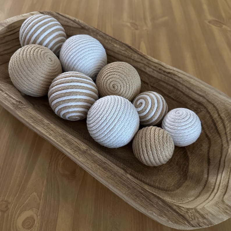 K&ouml;nnte beinhalten: Eine Sammlung dekorativer Kugeln in einer Holzschale. Die Kugeln bestehen aus gewebten Materialien in Beige, Wei&szlig; und Beige mit wei&szlig;en Streifen. Die Schale ist hellbraun und hat eine nat&uuml;rliche Holzmaserung. Die Kugeln sind unterschiedlich gro&szlig;.