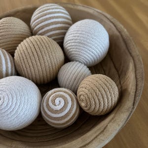 K&ouml;nnte beinhalten: Eine Holzschale gef&uuml;llt mit dekorativen Kugeln. Die Kugeln bestehen aus gewebtem Seil in Beige-, Creme- und Wei&szlig;t&ouml;nen. Einige Kugeln sind einfarbig, andere weisen Streifen- oder Spiralmuster auf.