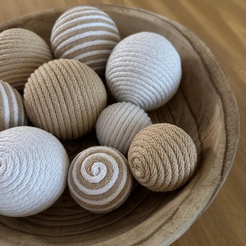 K&ouml;nnte beinhalten: Eine Holzschale gef&uuml;llt mit dekorativen Kugeln. Die Kugeln bestehen aus gewebtem Seil in Beige-, Creme- und Wei&szlig;t&ouml;nen. Einige Kugeln sind einfarbig, andere weisen Streifen- oder Spiralmuster auf.