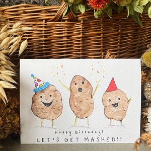 May include: A watercolour birthday card showing three potato characters wearing party hats and celebrating. The card has the text "Happy Birthday! LET'S GET MASHED!!". The potatoes are brown with green stems for arms and legs, and confetti is scattered above.