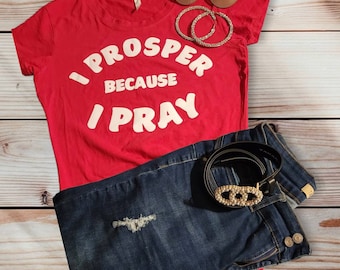 May include: Red t-shirt with white text that reads "I prosper because I pray". The shirt is paired with blue jeans and a brown belt with a gold buckle.