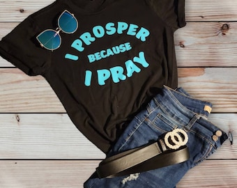 May include: Black t-shirt with the text "I Prosper Because I Pray" in blue lettering. The shirt is laid out on a wooden surface with a pair of sunglasses and a pair of blue jeans with a belt.