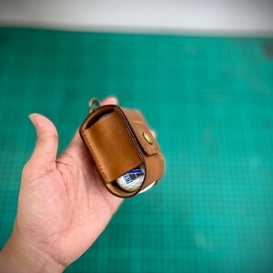 K&ouml;nnte beinhalten: Ein brauner Leder-Golfballhalter mit Druckknopfverschluss. Der Halter ist ge&ouml;ffnet und ein Golfball ist im Inneren sichtbar.