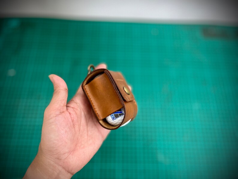 K&ouml;nnte beinhalten: Ein brauner Leder-Golfballhalter mit Druckknopfverschluss. Der Halter ist ge&ouml;ffnet und ein Golfball ist im Inneren sichtbar.