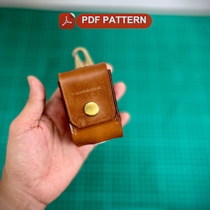 K&ouml;nnte beinhalten: Eine braune Ledertasche mit einem Messingknopfverschluss. Die Tasche wird in einer Hand gehalten.