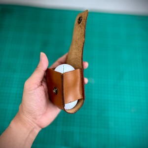 K&ouml;nnte beinhalten: Ein brauner Leder-Golfballhalter mit einem Golfball im Inneren. Der Halter ist ge&ouml;ffnet und der Golfball ist sichtbar.