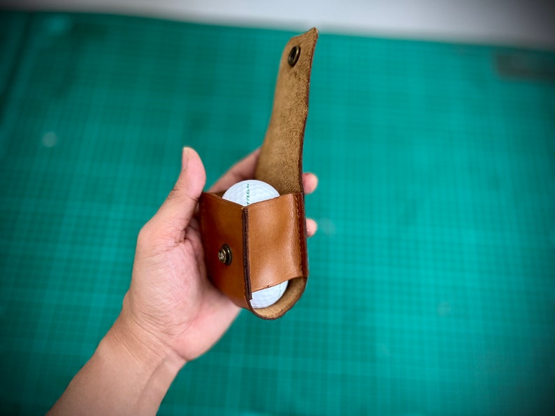 K&ouml;nnte beinhalten: Ein brauner Leder-Golfballhalter mit einem Golfball im Inneren. Der Halter ist ge&ouml;ffnet und der Golfball ist sichtbar.