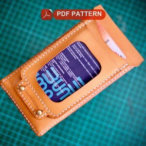 May include: A brown leather phone case with a stitched edge and a clear window for displaying a card. The case has a metal button and the text "HATTRICK" is visible on the front.