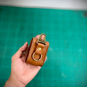 K&ouml;nnte beinhalten: Eine kleine braune Ledertasche mit einem Messingverschluss und einem Metallring. Die Tasche wird in einer Hand gehalten.