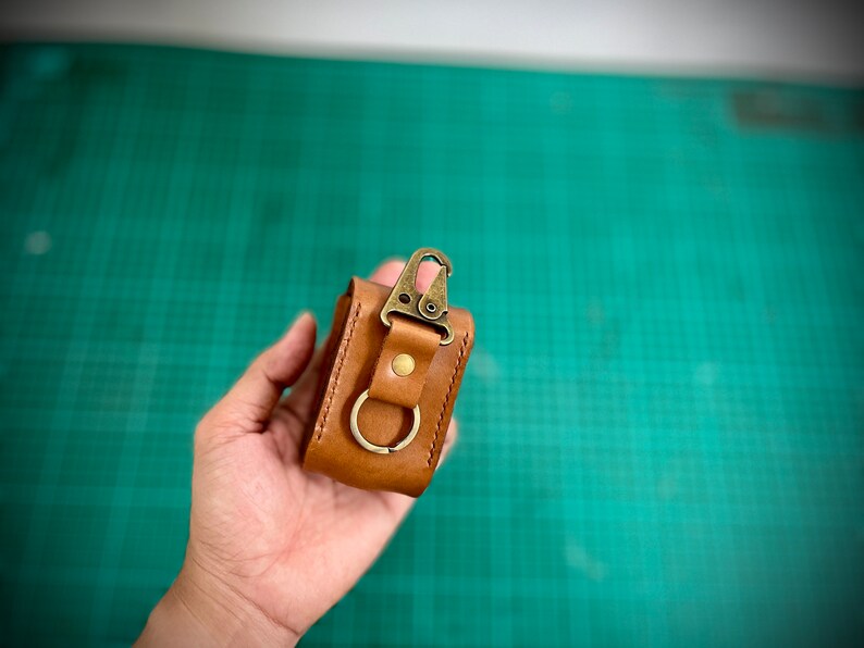 K&ouml;nnte beinhalten: Eine kleine braune Ledertasche mit einem Messingverschluss und einem Metallring. Die Tasche wird in einer Hand gehalten.