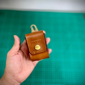 K&ouml;nnte beinhalten: Eine kleine braune Ledertasche mit einem Messingknopfverschluss. Die Tasche wird in einer Hand vor einem gr&uuml;nen Hintergrund gehalten.