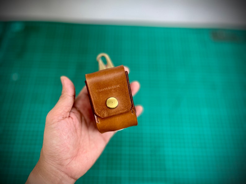 K&ouml;nnte beinhalten: Eine kleine braune Ledertasche mit einem Messingknopfverschluss. Die Tasche wird in einer Hand vor einem gr&uuml;nen Hintergrund gehalten.