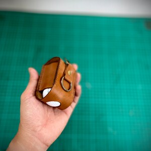 K&ouml;nnte beinhalten: Ein braunes Lederetui mit wei&szlig;em Innenfutter und einem Metallring. Das Etui wird in einer Hand gehalten.