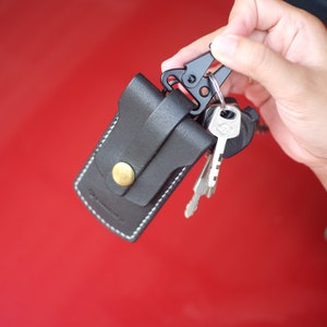 May include: A black leather key holder with a metal clip and a silver key attached. The key holder has a stitched edge and a small metal button.