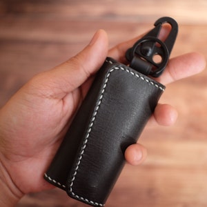 May include: A black leather key holder with white stitching and a black metal hook. The key holder is folded and held in a hand.