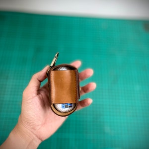 K&ouml;nnte beinhalten: Ein brauner Leder-Golfballhalter mit einem Metallclip. Der Halter ist ge&ouml;ffnet und ein wei&szlig;er Golfball ist im Inneren sichtbar.