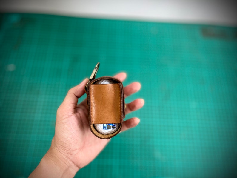 K&ouml;nnte beinhalten: Ein brauner Leder-Golfballhalter mit einem Metallclip. Der Halter ist ge&ouml;ffnet und ein wei&szlig;er Golfball ist im Inneren sichtbar.