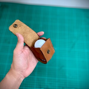 K&ouml;nnte beinhalten: Ein brauner Leder-Golfballhalter mit einem wei&szlig;en Golfball im Inneren. Der Halter hat einen Druckknopfverschluss und ist offen.