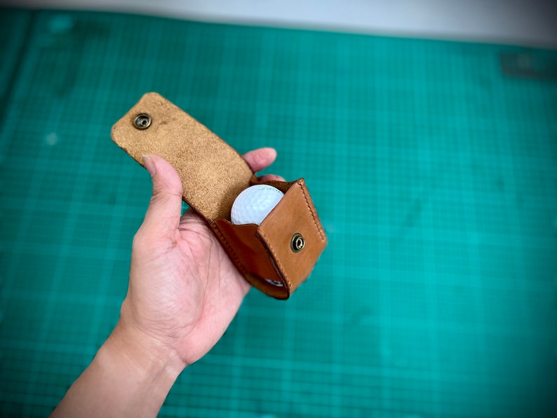 K&ouml;nnte beinhalten: Ein brauner Leder-Golfballhalter mit einem wei&szlig;en Golfball im Inneren. Der Halter hat einen Druckknopfverschluss und ist offen.