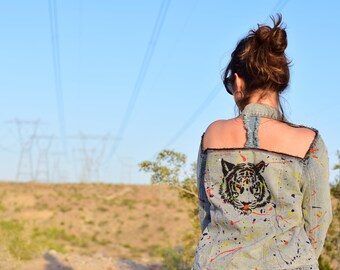 Könnte beinhalten: Eine Frau trägt eine hellblaue Jeansjacke mit einem schwarzen und weißen Tiger-Motiv auf dem Rücken. Die Jacke hat Farbschleier in verschiedenen Farben, darunter Rot, Gelb und Blau. Die Frau steht vor einer Wüstenlandschaft mit Stromleitungen im Hintergrund.