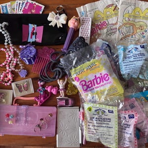 May include: A collection of vintage toys and accessories including a pink Barbie walkie-talkie, a black pencil case, a Miss Piggy Pez dispenser, various plastic jewelry, McDonald's Happy Meal bags, and small figurines scattered across a wooden surface.