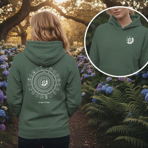 Könnte beinhalten: Ein dunkelgrüner Kapuzenpullover mit einem weißen kreisförmigen Design auf dem Rücken und einem kleineren Design auf der Vorderseite. Das Design auf der Rückseite zeigt ein detailliertes Muster mit Text am unteren Rand. Der Hoodie wird im Freien mit einem floralen Hintergrund gezeigt.
