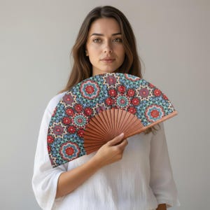 May include: A patterned hand fan with a wooden frame. The fan's fabric displays a repeating design of red, blue, and green floral motifs. The fan is open and held in front of a person, showcasing its decorative pattern.
