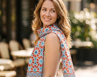 May include: A lightweight scarf featuring a geometric pattern in shades of terracotta orange and turquoise blue. The design consists of repeating star and polygon motifs with a decorative border along the edges, draped over the shoulders.