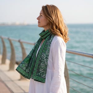 Può includere: Una sciarpa rettangolare caratterizzata da un intricato motivo verde e nero con dettagli floreali e geometrici bianchi. La sciarpa è drappeggiata attorno al collo, mettendo in risalto un bordo dettagliato e una stampa simmetrica ripetuta.