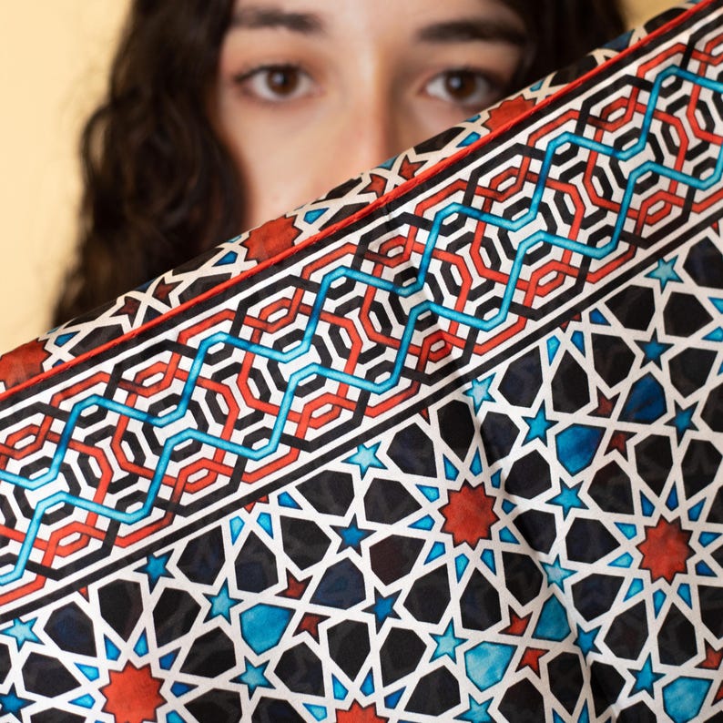 Large mulberry-silk scarf with Islamic geometry and Alhambra-inspired pattern, red border draped over a woman's shoulders.