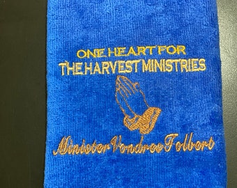 May include: A blue towel with gold embroidery that reads "One Heart for The Harvest Ministries" and "Minister Vondree Folbert". The embroidery also includes a pair of hands clasped in prayer.