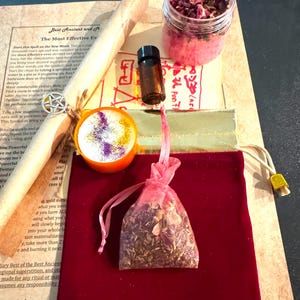 May include: An assortment of spiritual items, including a scroll with text, a small glass jar filled with pink granules and dried flowers, a small bottle, a small orange bowl with glitter, and a pink mesh bag filled with herbs, all arranged on a red velvet surface.