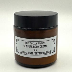 May include: A brown glass jar with a black lid. The jar has a white label with the text "BEST SPELLS MAGICK CONJURE BODY CREAM Silk LLAMA CLIENTE/BETTER BUSINES". The jar is sitting on a white surface.