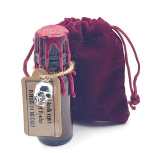 May include: A small brown glass bottle with a red wax seal and a tag that reads "Spells Magick Art Powder  POWDER OF THE DEAD". The bottle is in a small red velvet bag with a drawstring.