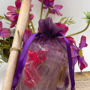 May include: A purple organza bag filled with various items for a protection spell. The bag contains a red string, a silver pentagram charm, a small bottle of oil, and a rolled up piece of paper. The bag is surrounded by pink and purple flowers.