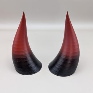 Könnte beinhalten: Ein Paar Teufelshörner mit Farbverlauf. Die Hörner wechseln von tiefem Rot an den Spitzen zu Schwarz an der Basis. Die Hörner haben eine gebogene Form und eine strukturierte Oberfläche.