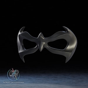Könnte beinhalten: Eine dunkelgraue Maske mit spitzem Mittelteil und gebogenen Kanten. Die Maske hat Augenöffnungen und ist wahrscheinlich für Kostüm- oder Cosplay-Zwecke gedacht. Das Bild hat einen dunklen Hintergrund und die Maske steht im Mittelpunkt.