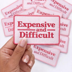 May include: A collection of pink and white embroidered patches with the text "Expensive and Difficult" in red. One patch is held in a hand, with several others scattered in the background. The patches have a rectangular shape.