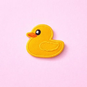 May include: A yellow embroidered duck patch with an orange beak and a black eye. The patch is on a pink background. The duck is a classic cartoon design.