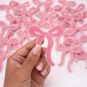 May include: A collection of pink embroidered bow patches featuring a stitched outline and two long, flowing tails. Each patch is crafted with a dense thread texture, showcasing a consistent shape and vibrant pink color throughout the design.