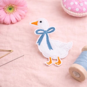 May include: A white goose-shaped felt applique with a blue ribbon around its neck, orange feet, and a yellow beak. The applique is on a pink fabric surface, with a pink flower and sewing notions in the background.