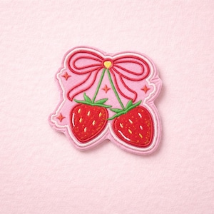 May include: A pink patch featuring two red strawberries with green stems, hanging from a pink bow. The strawberries have white outlines and small white seeds. The patch has a pink background with star accents.