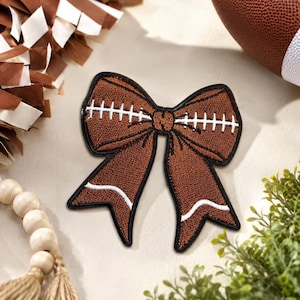 May include: A brown embroidered patch in the shape of a bow, featuring white stitched details that mimic the laces and stripes of an American football. The patch has a black border and a textured surface, designed to resemble the leather of a ball.