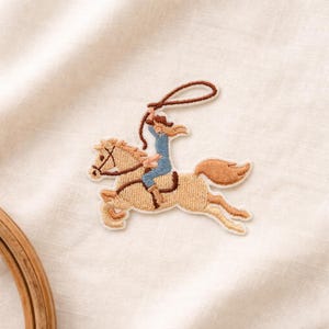 May include: Embroidered patch featuring a cowboy riding a horse. The horse is tan with a brown mane and tail, and the cowboy is wearing a blue outfit and a brown hat. The cowboy is holding a lasso. The patch is on a white background.