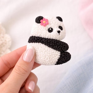 May include: A handmade panda bear pin with a white body, black accents, and a pink flower. The panda has a textured, fuzzy appearance. The pin is held by a person.