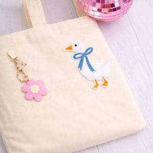 May include: Cream canvas tote bag featuring a white duck patch with a blue bow. A pink flower keyring hangs from the bag. A small pink disco ball is in the background. The bag has a simple design.