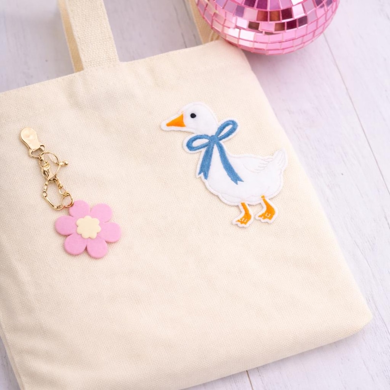 May include: Cream canvas tote bag featuring a white duck patch with a blue bow. A pink flower keyring hangs from the bag. A small pink disco ball is in the background. The bag has a simple design.