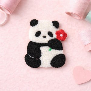 May include: A handmade panda bear applique with black and white fur, holding a red flower. The panda has black eyes and ears. The applique is made of textured fabric and is set against a pink background with spools of thread.