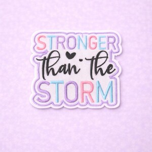 May include: A white patch with the words "STRONGER than the STORM" in a mix of colors and fonts. The patch has a purple border and is set against a light purple background.
