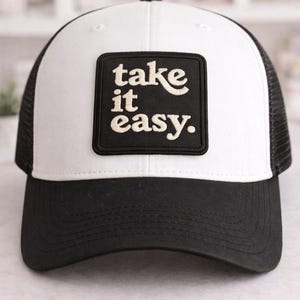May include: A black and white trucker hat with a white front panel and a black mesh back. The hat features a black square patch with the words "take it easy" in a white, cursive font.